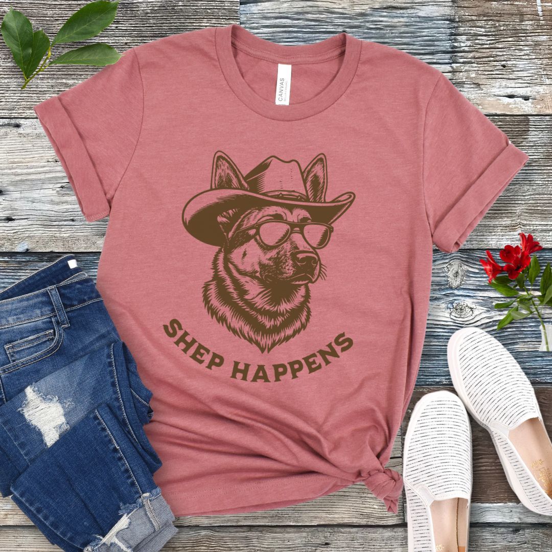 Mauve t-shirt with a graphic of a German Shepherd dog wearing sunglasses and a cowboy hat, with text 'SHEP HAPPENS', displayed on a wooden surface with jeans and shoes.