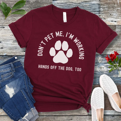 A maroon T-shirt with the text 'DON'T PET ME, I'M WORKING. HANDS OFF THE DOG, TOO' and a paw print design, displayed on a wooden surface with a pair of jeans and white shoes in the background.