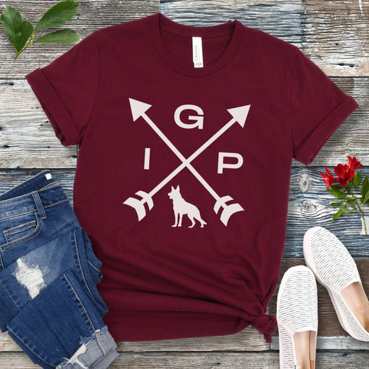 Maroon IGP dog t-shirt with a graphic of crossed arrows, the letters IGP, and silhouette of a German Shepherd Dog, displayed on a wooden surface with a pair of jeans and white shoes in the background.