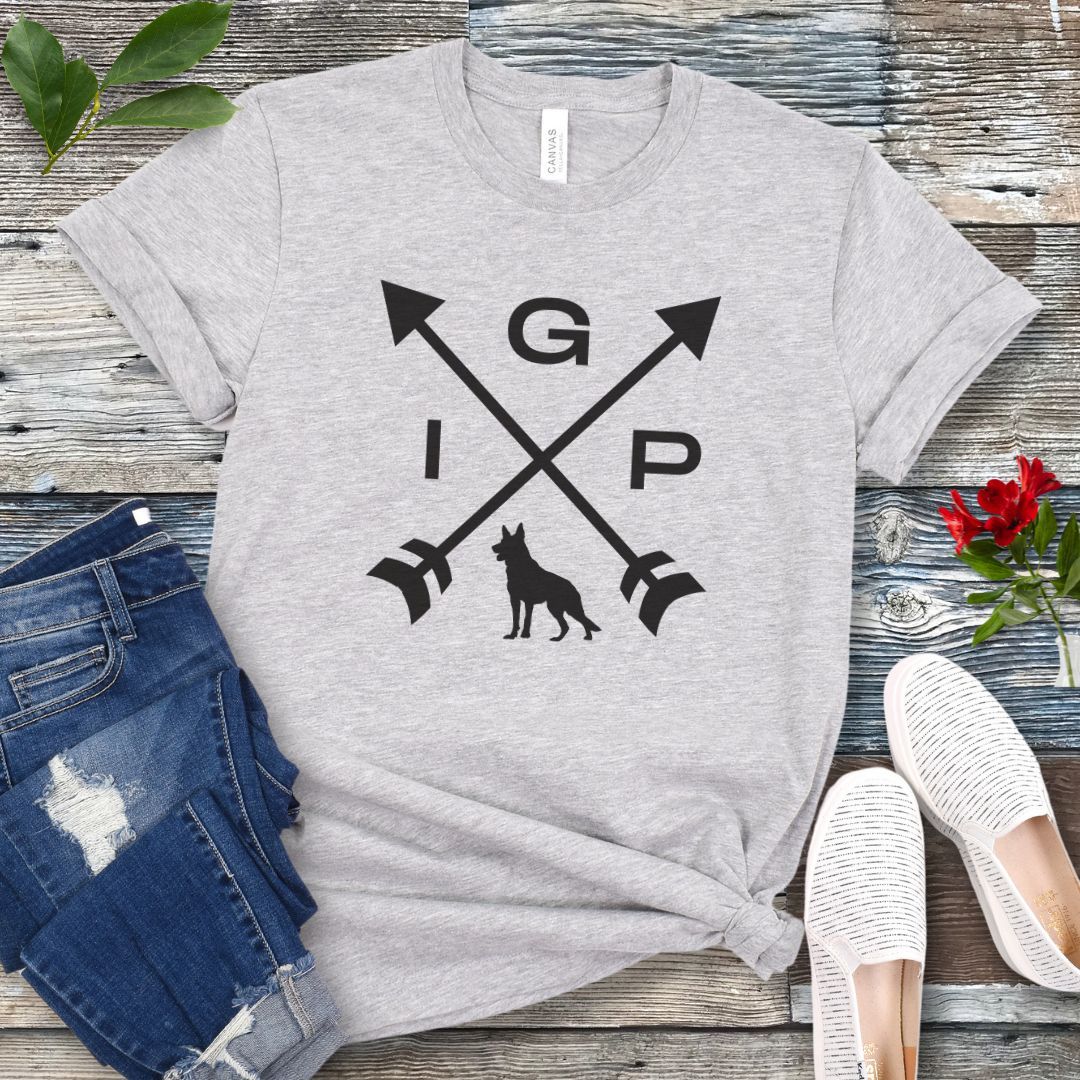 Grey IGP dog t-shirt with a graphic of crossed arrows, the letters IGP, and silhouette of a German Shepherd Dog, displayed on a wooden surface with a pair of jeans and white shoes in the background.