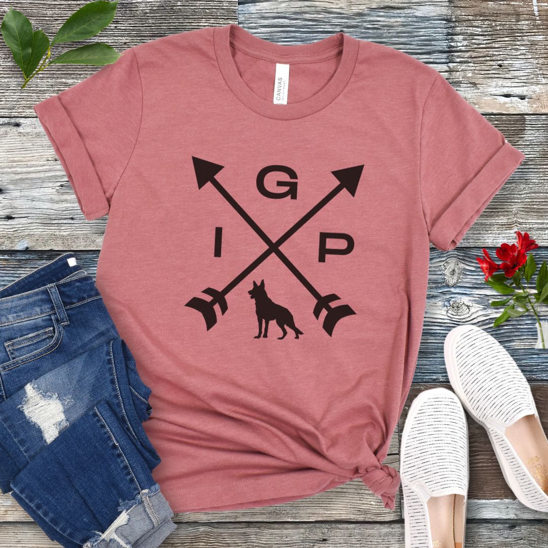 Mauve IGP dog t-shirt with a graphic of crossed arrows, the letters IGP, and silhouette of a German Shepherd Dog, displayed on a wooden surface with a pair of jeans and white shoes in the background.