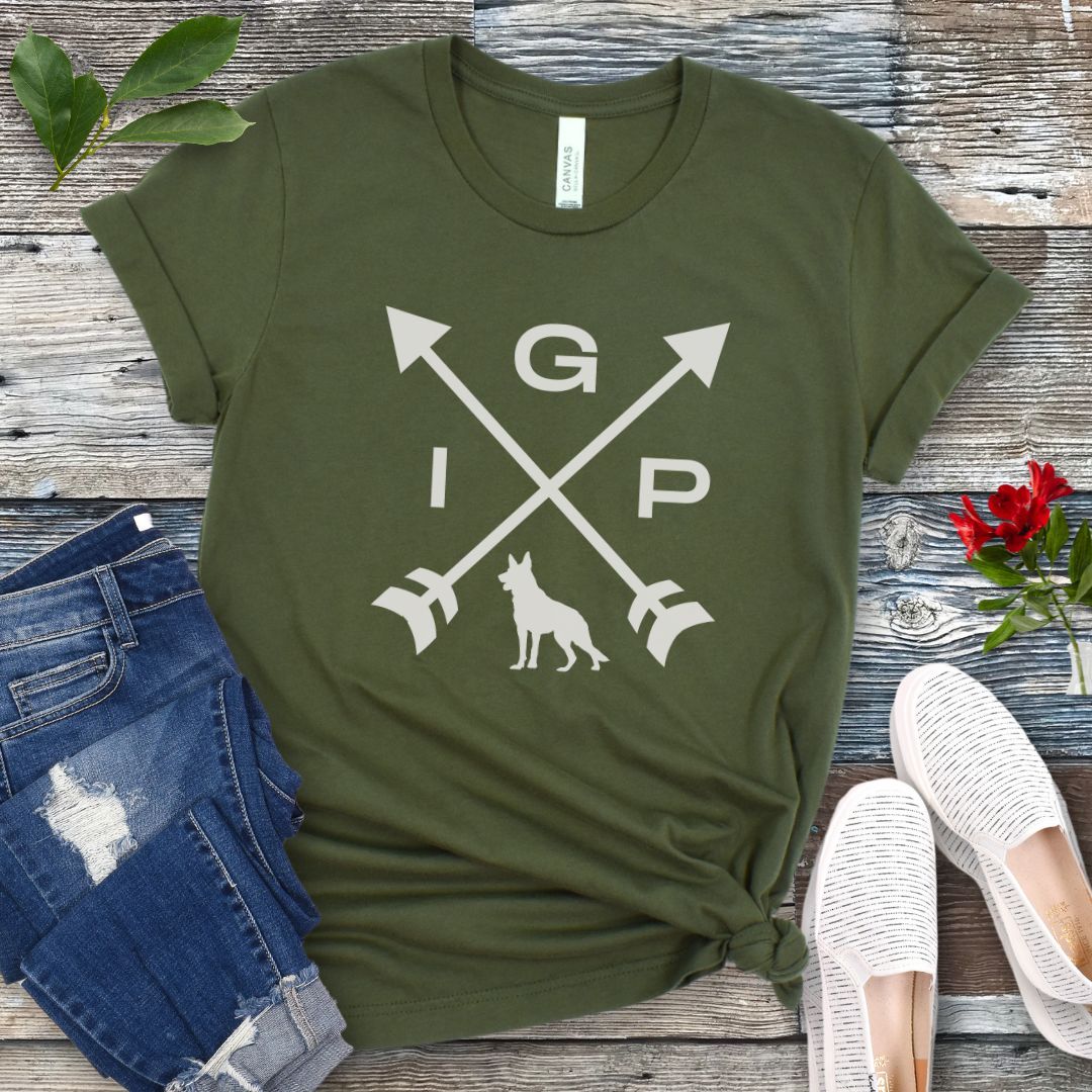 Military green IGP dog t-shirt with a graphic of crossed arrows, the letters IGP, and silhouette of a German Shepherd Dog, displayed on a wooden surface with a pair of jeans and white shoes in the background.