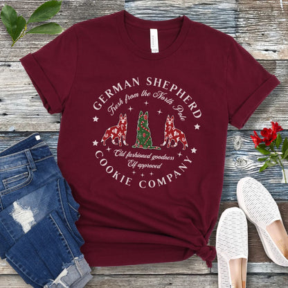 Maroon t-shirt with German Shepherd-shaped Christmas cookies and text  'German Shepherd Cookie Company', presented on a wooden surface with jeans and shoes.