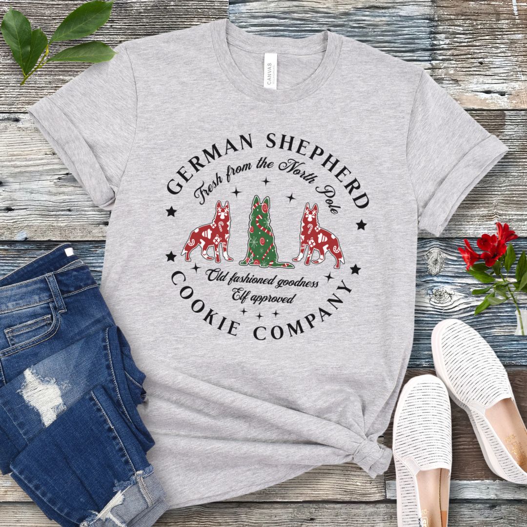 Grey t-shirt with German Shepherd-shaped Christmas cookies and text  'German Shepherd Cookie Company', presented on a wooden surface with jeans and shoes.