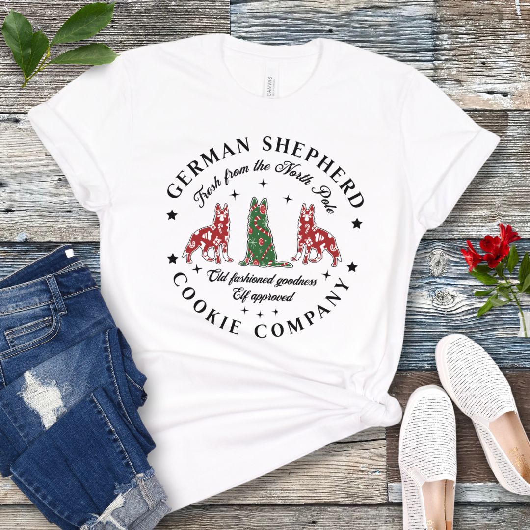 White t-shirt with German Shepherd-shaped Christmas cookies and text  'German Shepherd Cookie Company', presented on a wooden surface with jeans and shoes.