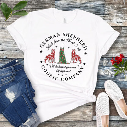 White t-shirt with German Shepherd-shaped Christmas cookies and text  'German Shepherd Cookie Company', presented on a wooden surface with jeans and shoes.