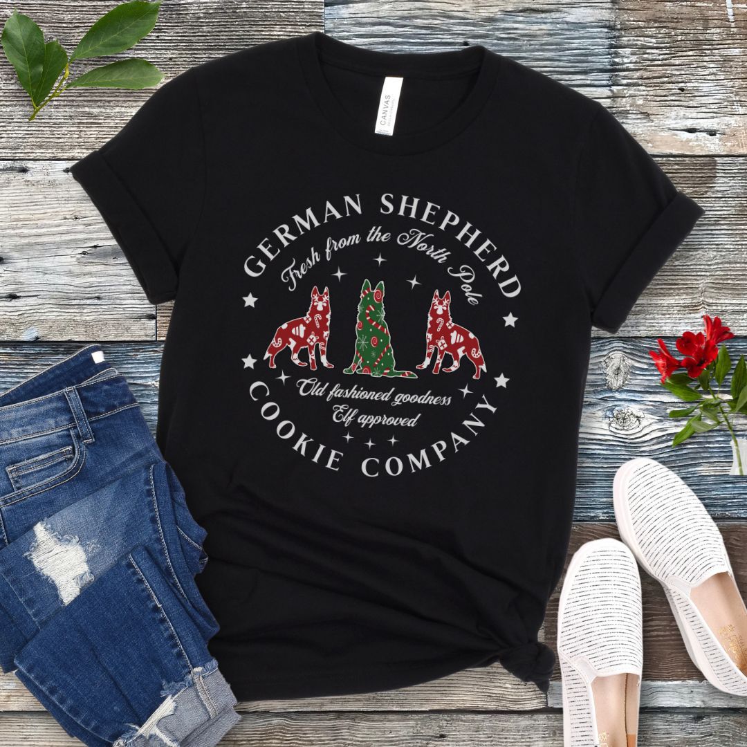 Black t-shirt with German Shepherd-shaped Christmas cookies and text  'German Shepherd Cookie Company', presented on a wooden surface with jeans and shoes.
