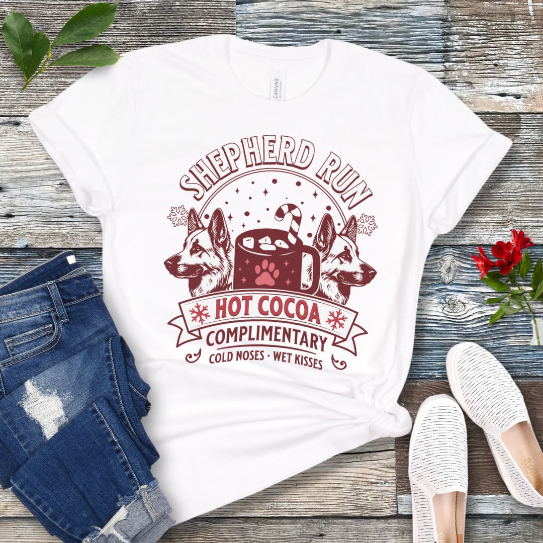 White t-shirt with German Shepherds, hot cocoa, candy cane and snowflakes, with the text 'Shepherd Run Hot Cocoa Company',  presented on a wooden surface with jeans and shoes.