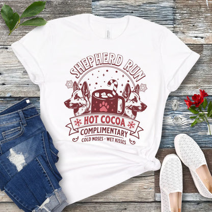 White t-shirt with German Shepherds, hot cocoa, candy cane and snowflakes, with the text 'Shepherd Run Hot Cocoa Company',  presented on a wooden surface with jeans and shoes.
