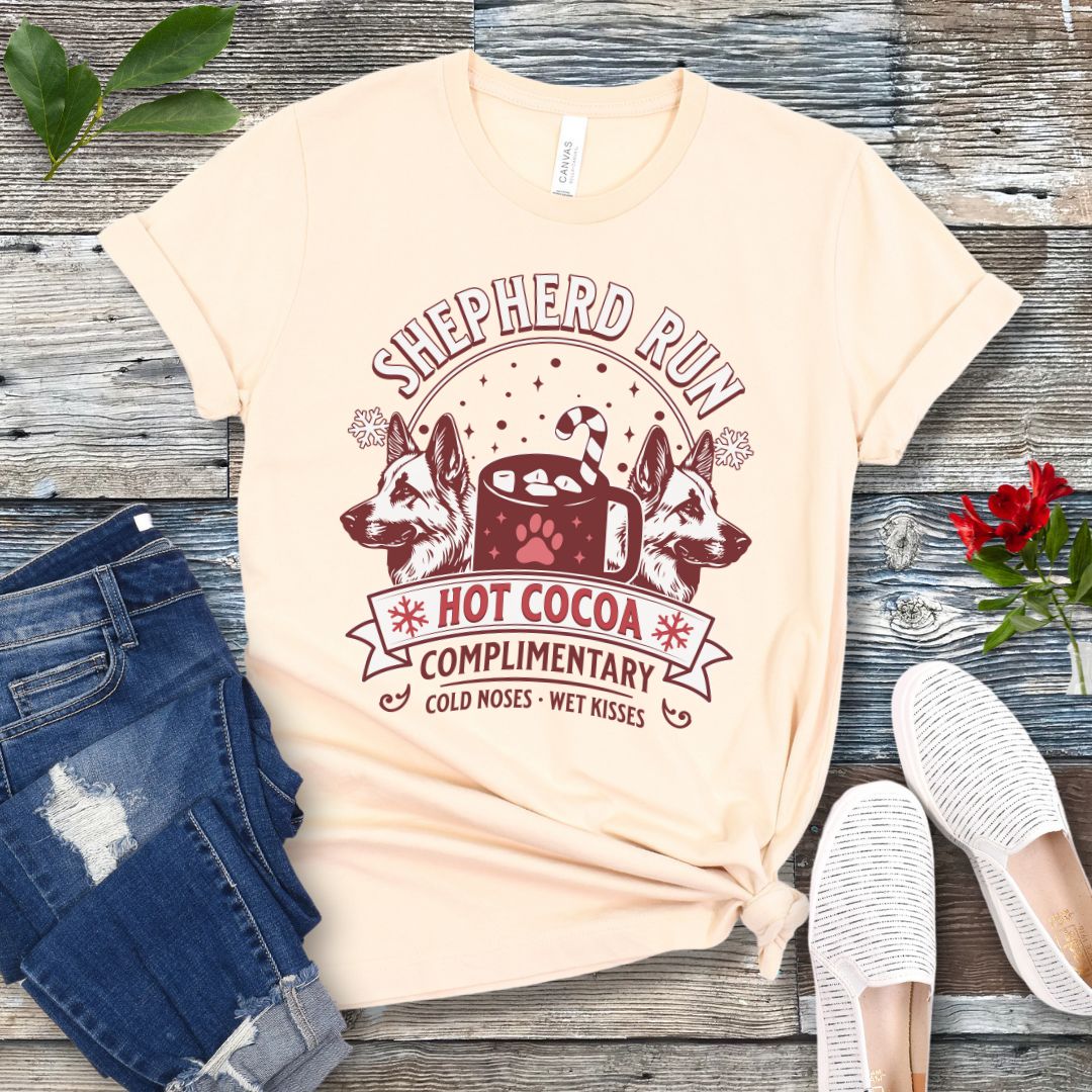 Beige t-shirt with German Shepherds, hot cocoa, candy cane and snowflakes, with the text 'Shepherd Run Hot Cocoa Company',  presented on a wooden surface with jeans and shoes.