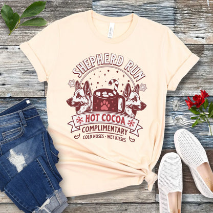 Beige t-shirt with German Shepherds, hot cocoa, candy cane and snowflakes, with the text 'Shepherd Run Hot Cocoa Company',  presented on a wooden surface with jeans and shoes.