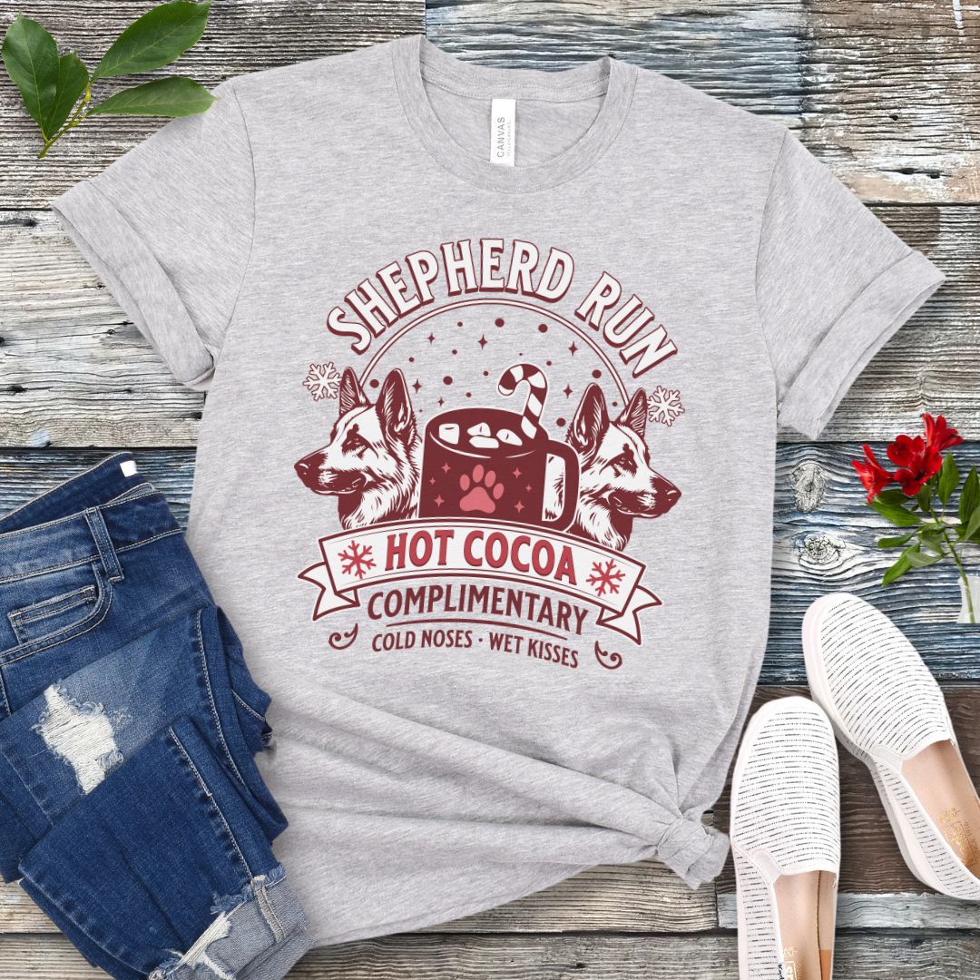 Grey t-shirt with German Shepherds, hot cocoa, candy cane and snowflakes, with the text 'Shepherd Run Hot Cocoa Company',  presented on a wooden surface with jeans and shoes.