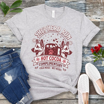 Grey t-shirt with German Shepherds, hot cocoa, candy cane and snowflakes, with the text 'Shepherd Run Hot Cocoa Company',  presented on a wooden surface with jeans and shoes.