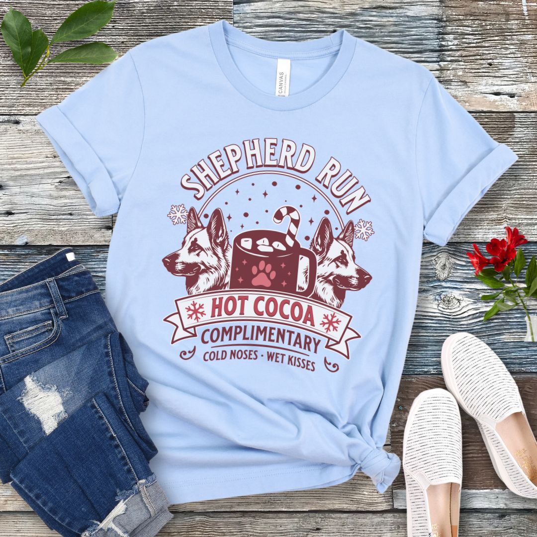Light blue t-shirt with German Shepherds, hot cocoa, candy cane and snowflakes, with the text 'Shepherd Run Hot Cocoa Company',  presented on a wooden surface with jeans and shoes.