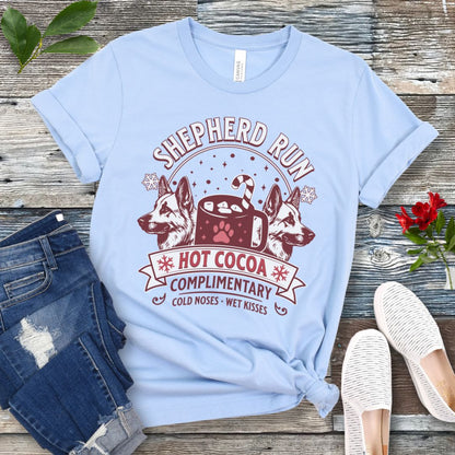 Light blue t-shirt with German Shepherds, hot cocoa, candy cane and snowflakes, with the text 'Shepherd Run Hot Cocoa Company',  presented on a wooden surface with jeans and shoes.