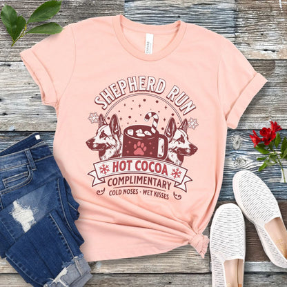 Peach t-shirt with German Shepherds, hot cocoa, candy cane and snowflakes, with the text 'Shepherd Run Hot Cocoa Company',  presented on a wooden surface with jeans and shoes.