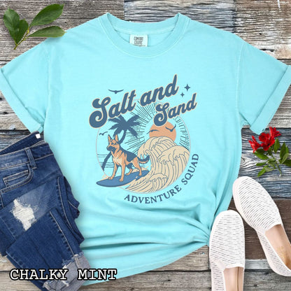Mint Comfort Colors t-shirt for German Shepherd lovers with beach-themed graphic of a surfing GSD, waves and sunset, and the text 'Salt and Sand Adventure Squad' in retro lettering, shown on a wooden surface with accessories.