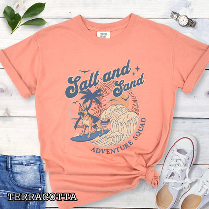 Orange Comfort Colors t-shirt for German Shepherd lovers with beach-themed graphic of a surfing GSD, waves and sunset, and the text 'Salt and Sand Adventure Squad' in retro lettering, shown on a wooden surface with accessories.