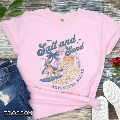 Pink Comfort Colors t-shirt for German Shepherd lovers with beach-themed graphic of a surfing GSD, waves and sunset, and the text 'Salt and Sand Adventure Squad' in retro lettering, shown on a wooden surface with accessories.