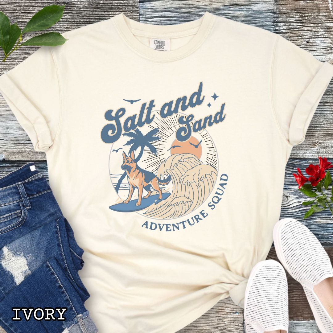 Beige Comfort Colors t-shirt for German Shepherd lovers with beach-themed graphic of a surfing GSD, waves and sunset, and the text 'Salt and Sand Adventure Squad' in retro lettering, shown on a wooden surface with accessories.