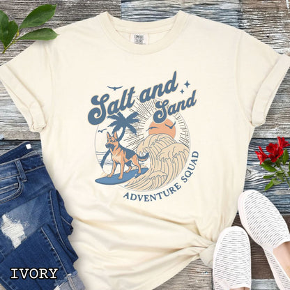 Beige Comfort Colors t-shirt for German Shepherd lovers with beach-themed graphic of a surfing GSD, waves and sunset, and the text 'Salt and Sand Adventure Squad' in retro lettering, shown on a wooden surface with accessories.