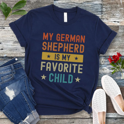 Navy blue t-shirt with colorful text 'My German Shepherd is my Favorite Child' on a wooden surface with jeans and shoes.