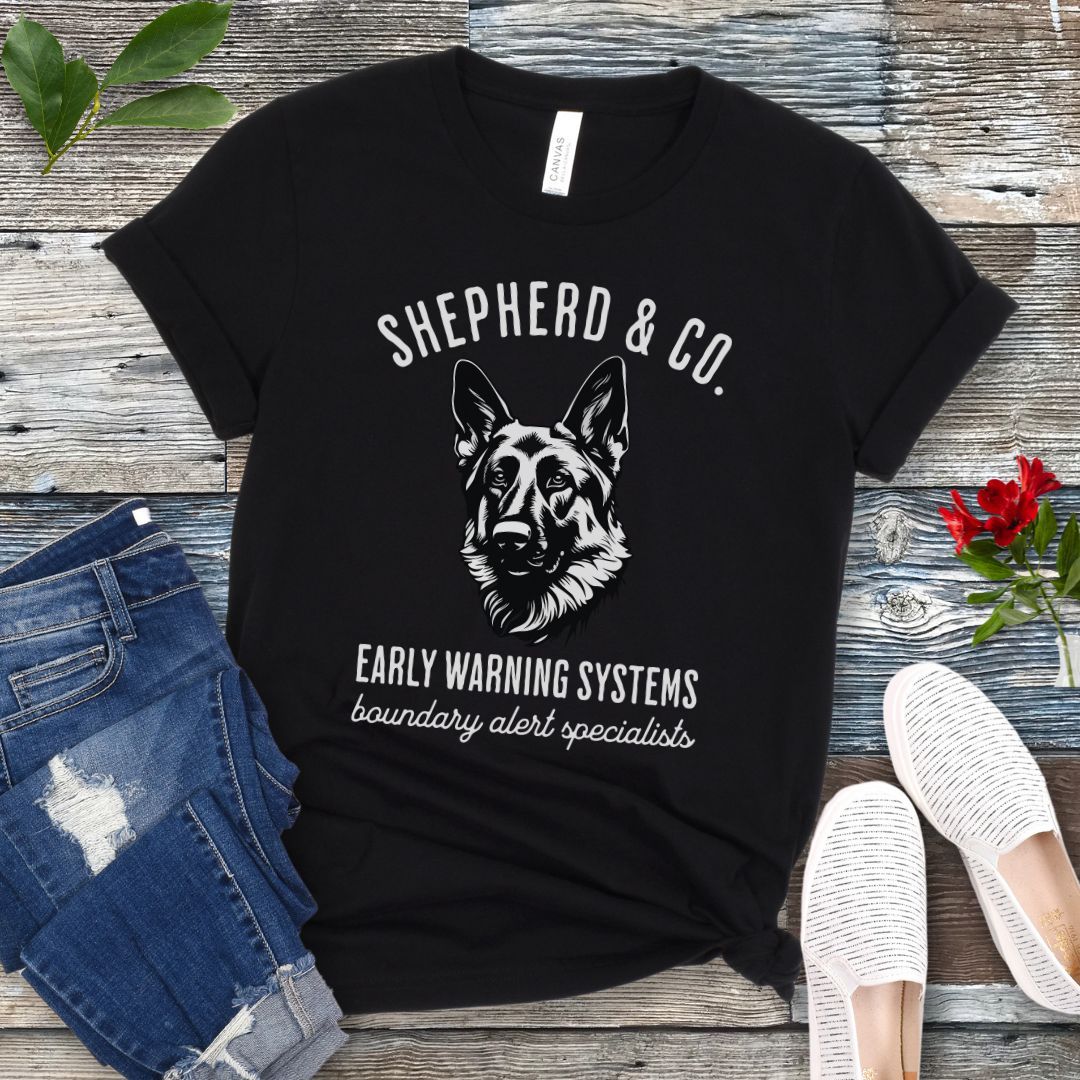 Black t-shirt with a German Shepherd dog graphic and 'Shepherd and Co. Early Warning Systems' text, presented on a wooden surface with jeans and shoes.