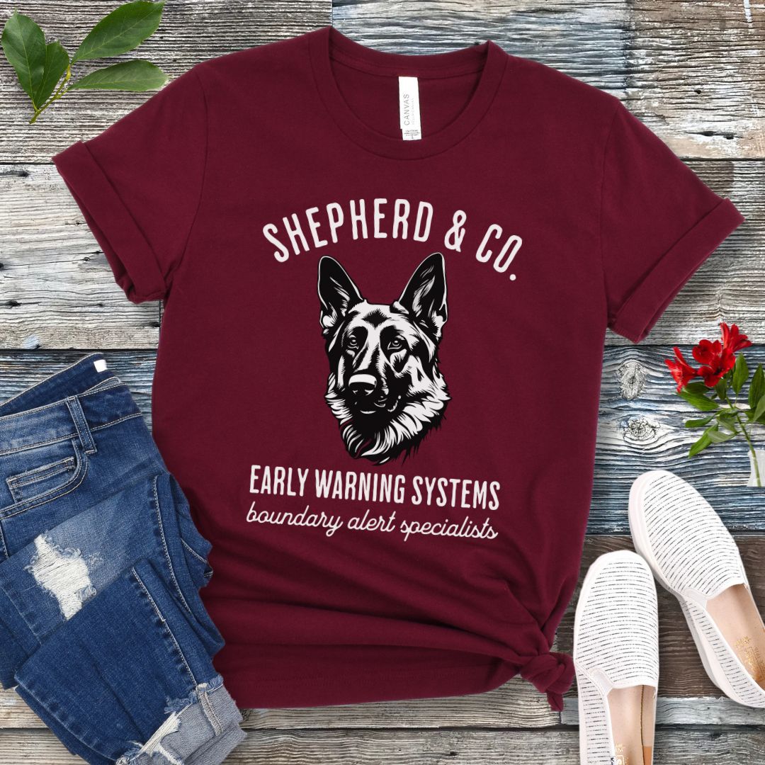 Maroon t-shirt with a German Shepherd dog graphic and 'Shepherd and Co. Early Warning Systems' text, presented on a wooden surface with jeans and shoes.