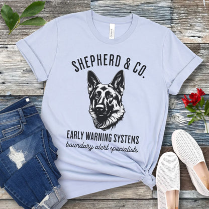 Light blue t-shirt with a German Shepherd dog graphic and 'Shepherd and Co. Early Warning Systems' text, presented on a wooden surface with jeans and shoes.