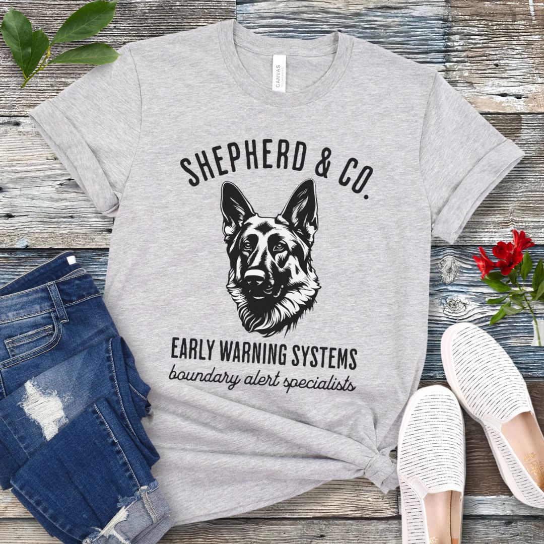Gray t-shirt with a German Shepherd dog graphic and 'Shepherd and Co. Early Warning Systems' text, presented on a wooden surface with jeans and shoes.