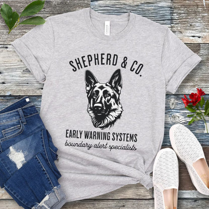 Gray t-shirt with a German Shepherd dog graphic and 'Shepherd and Co. Early Warning Systems' text, presented on a wooden surface with jeans and shoes.