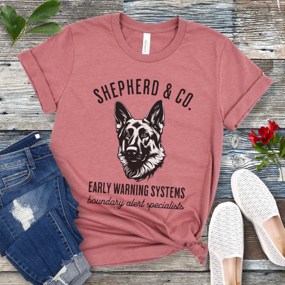 Mauve t-shirt with a German Shepherd dog graphic and 'Shepherd and Co. Early Warning Systems' text, presented on a wooden surface with jeans and shoes.