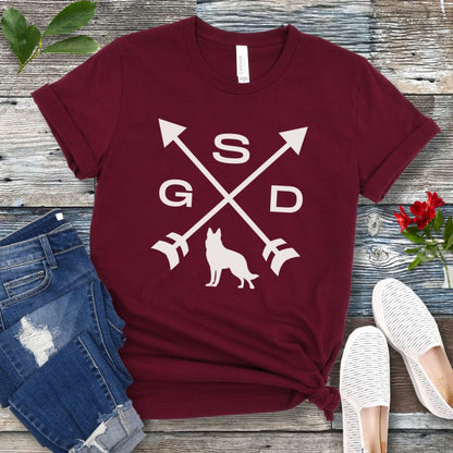 Maroon t-shirt with a graphic of crossed arrows, the letters GSD, and silhouette of a German Shepherd Dog in the center, displayed on a wooden surface with a pair of jeans and white shoes in the background.