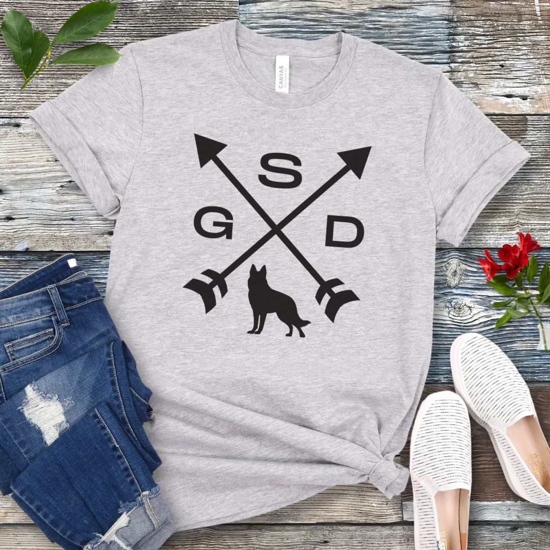 Grey t-shirt with a graphic of crossed arrows, the letters GSD, and silhouette of a German Shepherd Dog in the center, displayed on a wooden surface with a pair of jeans and white shoes in the background.
