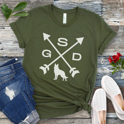 Military green t-shirt with a graphic of crossed arrows, the letters GSD, and silhouette of a German Shepherd Dog in the center, displayed on a wooden surface with a pair of jeans and white shoes in the background.