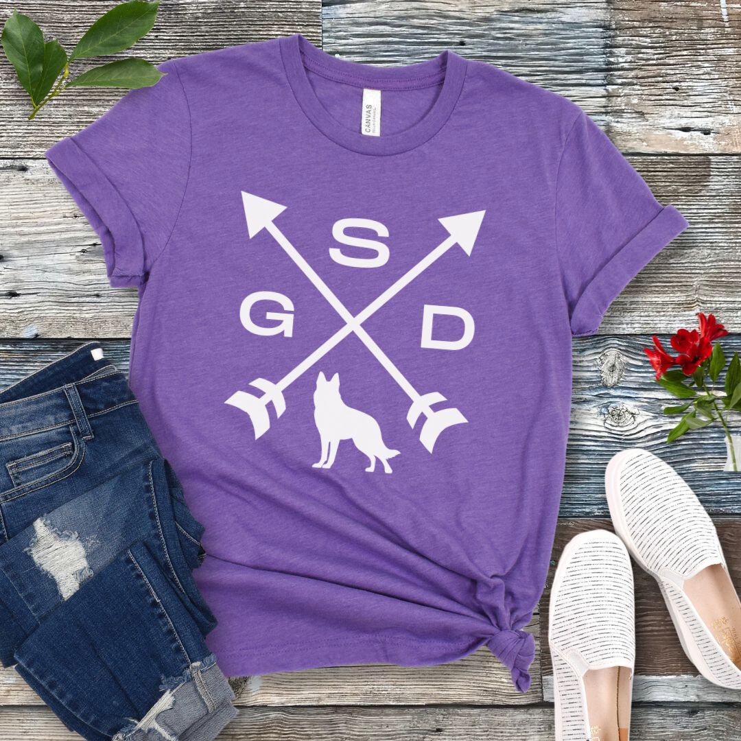 Purple t-shirt with a graphic of crossed arrows, the letters GSD, and silhouette of a German Shepherd Dog in the center, displayed on a wooden surface with a pair of jeans and white shoes in the background.