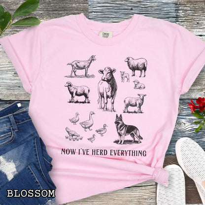 Pink Comfort Colors t-shirt for German Shepherd lovers interested in breed working history, with vintage farm animal illustrations and a German Shepherd dog, with the humorous text 'Now I've Herd Everything', shown on a wooden surface with denim jeans and flowers.