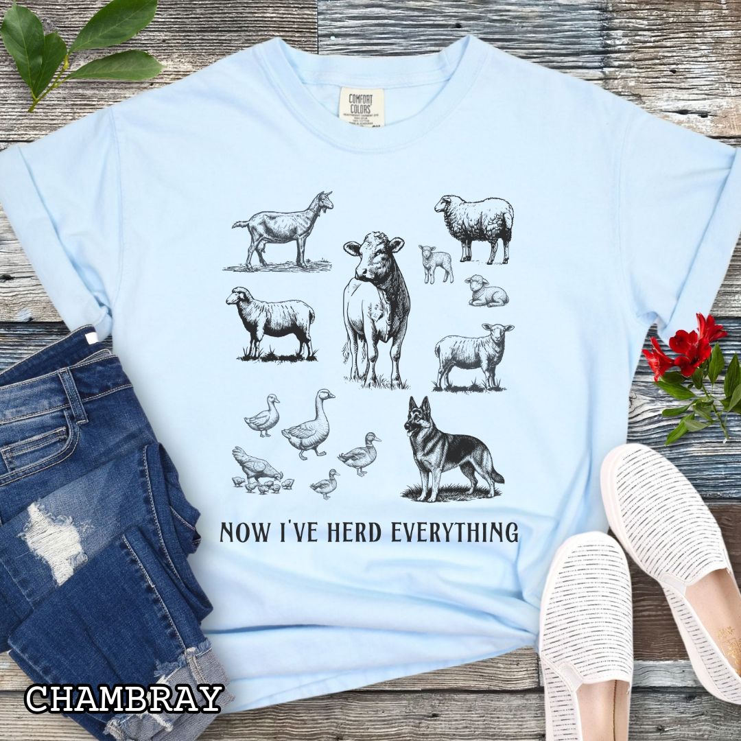 Light blue Comfort Colors t-shirt for German Shepherd lovers interested in breed working history, with vintage farm animal illustrations and a German Shepherd dog, with the humorous text 'Now I've Herd Everything', shown on a wooden surface with denim jeans and flowers.