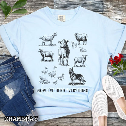 Light blue Comfort Colors t-shirt for German Shepherd lovers interested in breed working history, with vintage farm animal illustrations and a German Shepherd dog, with the humorous text 'Now I've Herd Everything', shown on a wooden surface with denim jeans and flowers.