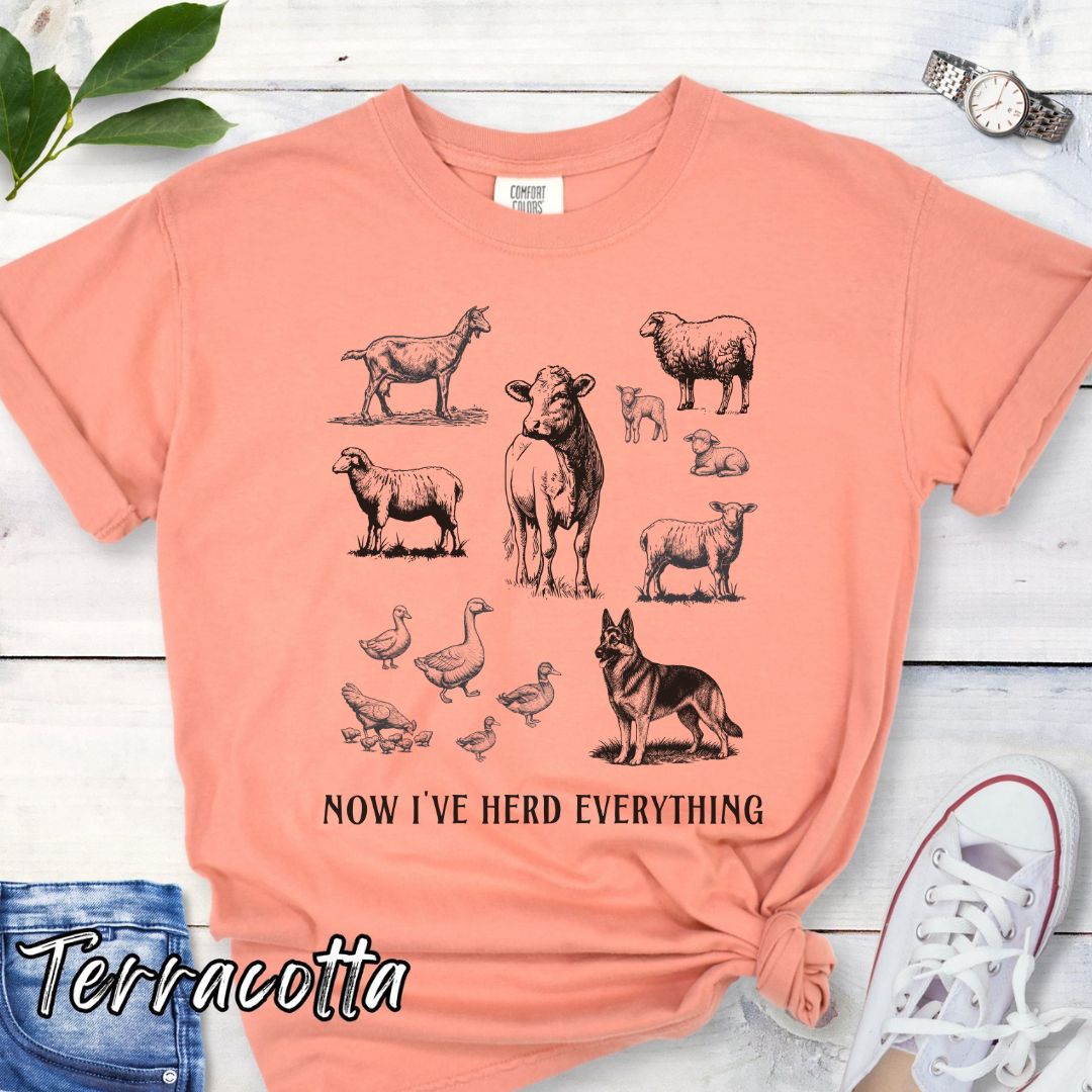Orange Comfort Colors t-shirt for German Shepherd lovers interested in breed working history, with vintage farm animal illustrations and a German Shepherd dog, with the humorous text 'Now I've Herd Everything', shown on a wooden surface with denim jeans and flowers.