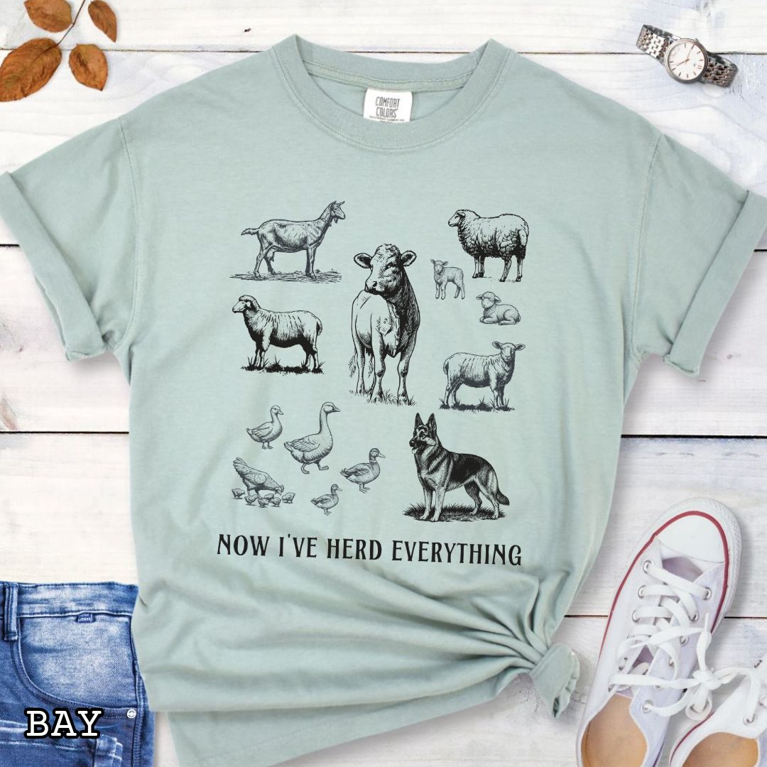 Pale green Comfort Colors t-shirt for German Shepherd lovers interested in breed working history, with vintage farm animal illustrations and a German Shepherd dog, with the humorous text 'Now I've Herd Everything', shown on a wooden surface with denim jeans and flowers.