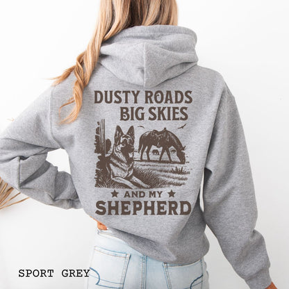 Back view of grey hoodie featuring a Western-style design with grazing horse, rustic fence, and a German Shepherd illustration; text reads “Dusty Roads, Big Skies, and My Shepherd.”