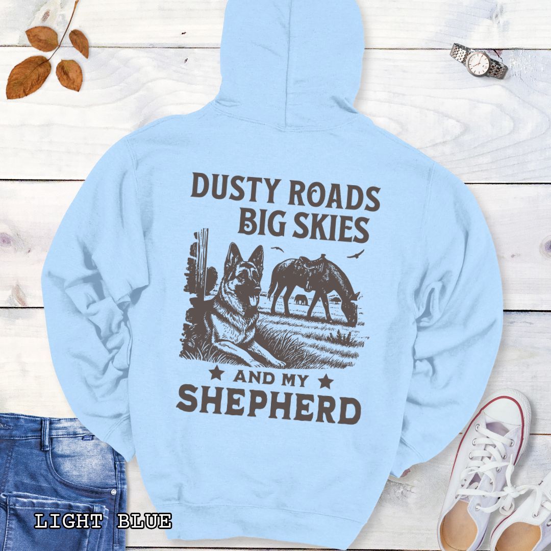 Back view of light blue hoodie featuring a Western-style design with grazing horse, rustic fence, and a German Shepherd illustration; text reads “Dusty Roads, Big Skies, and My Shepherd.”