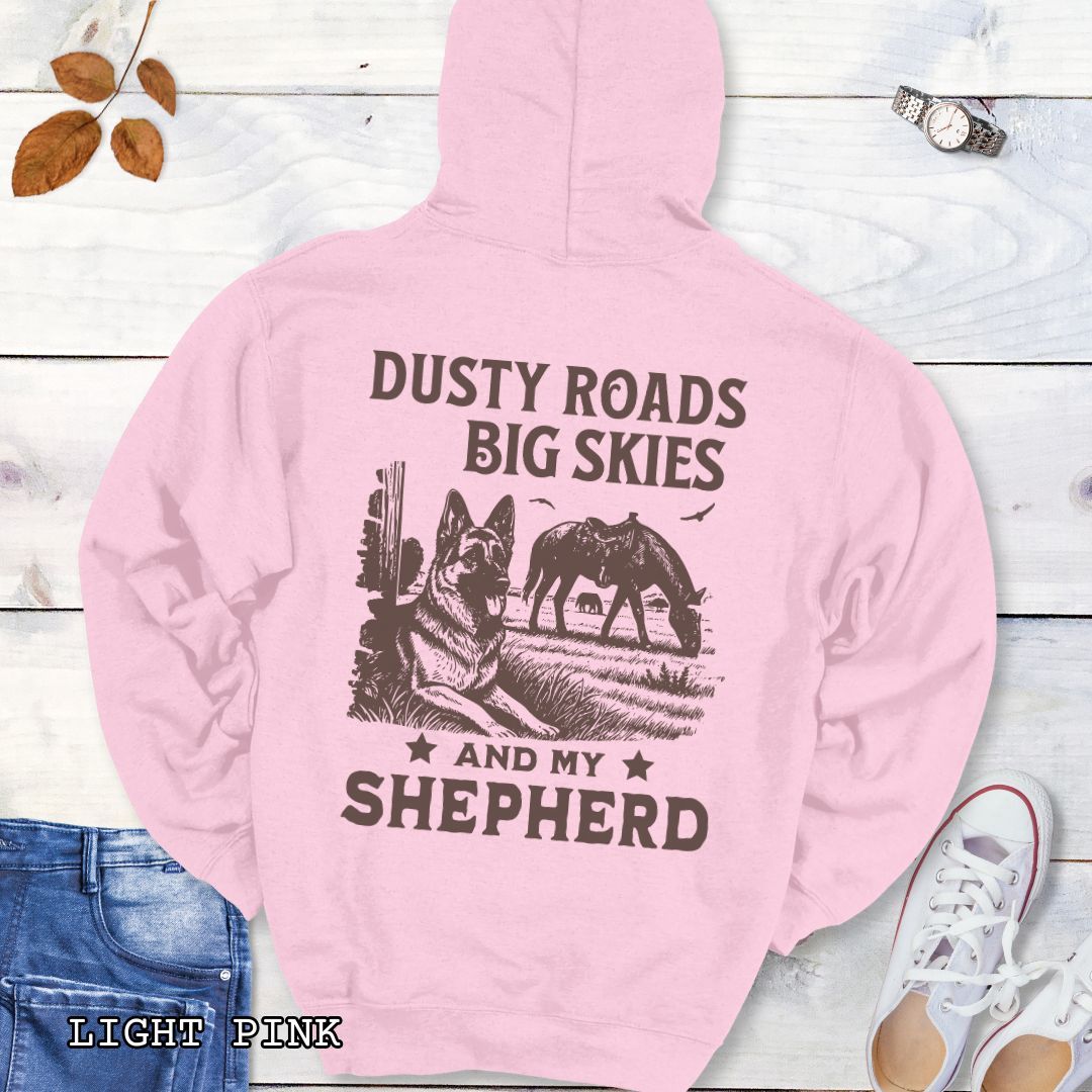 Back view of light pink hoodie featuring a Western-style design with grazing horse, rustic fence, and a German Shepherd illustration; text reads “Dusty Roads, Big Skies, and My Shepherd.”