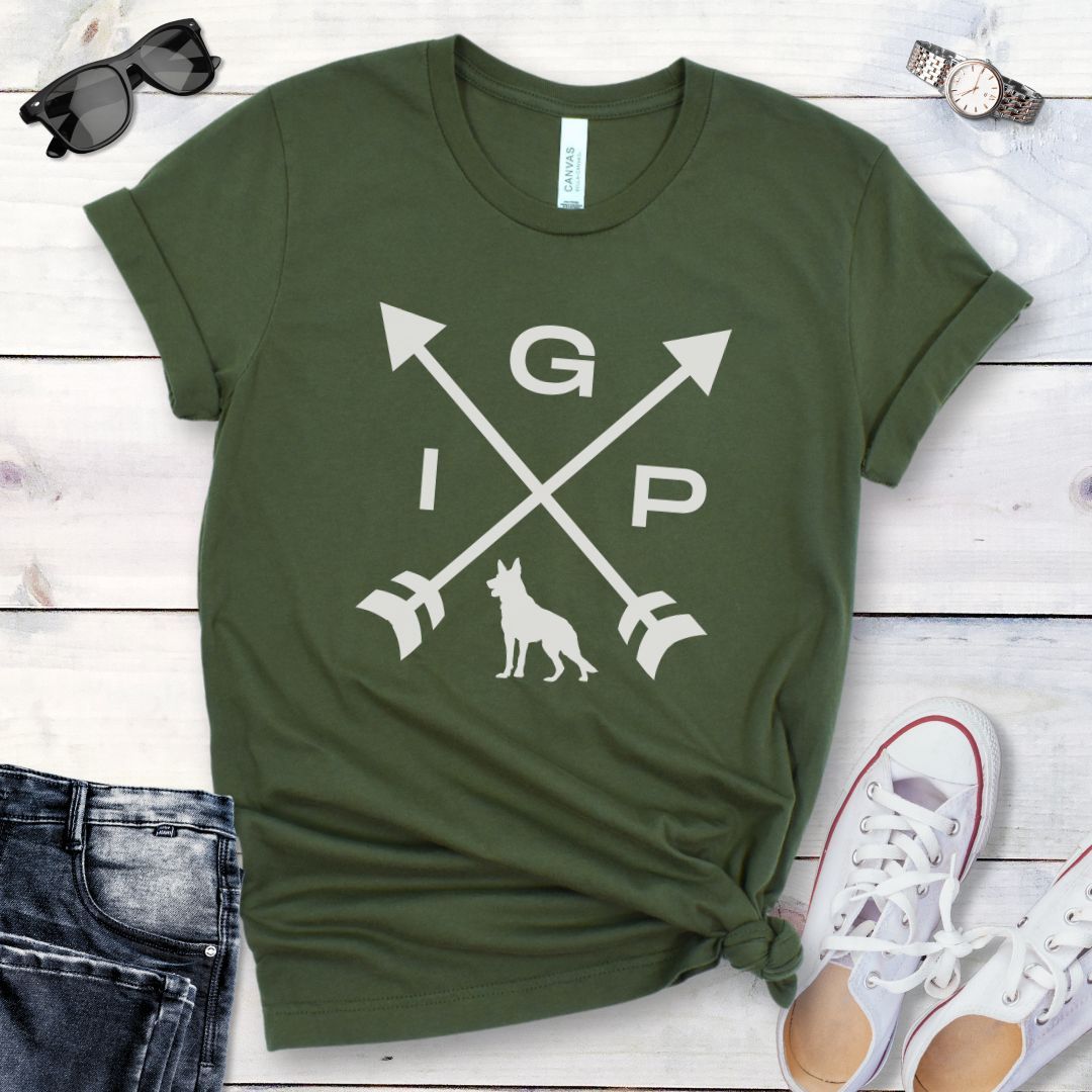 Military green IGP dog t-shirt with a graphic of crossed arrows, the letters IGP, and silhouette of a German Shepherd Dog, displayed on a wooden surface with a pair of jeans and white shoes in the background.