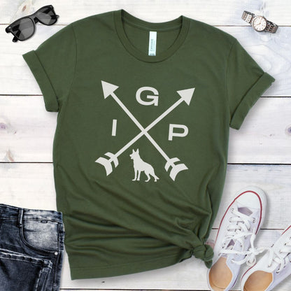 Military green IGP dog t-shirt with a graphic of crossed arrows, the letters IGP, and silhouette of a German Shepherd Dog, displayed on a wooden surface with a pair of jeans and white shoes in the background.
