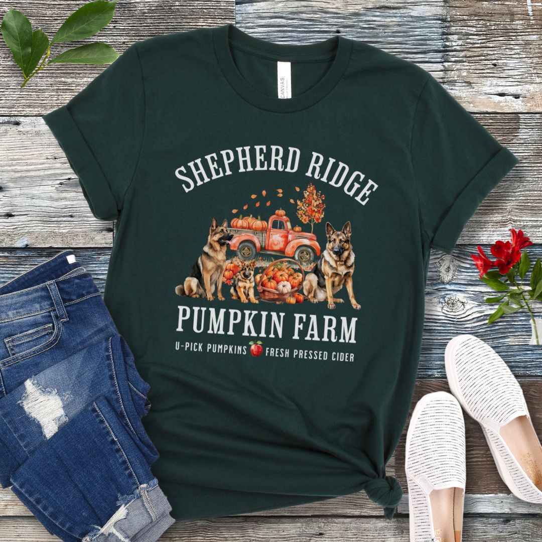 Forest green t-shirt with watercolor graphic of German Shepherds, a pumpkins truck and fall leaves, with the text 'Shepherd Ridge Pumpkin Farm', presented on a wooden surface with jeans and shoes.