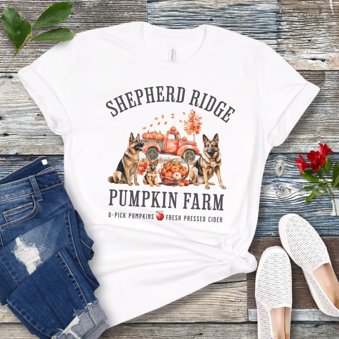White t-shirt with watercolor graphic of German Shepherds, a pumpkins truck and fall leaves, with the text 'Shepherd Ridge Pumpkin Farm', presented on a wooden surface with jeans and shoes.