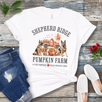 White t-shirt with watercolor graphic of German Shepherds, a pumpkins truck and fall leaves, with the text 'Shepherd Ridge Pumpkin Farm', presented on a wooden surface with jeans and shoes.