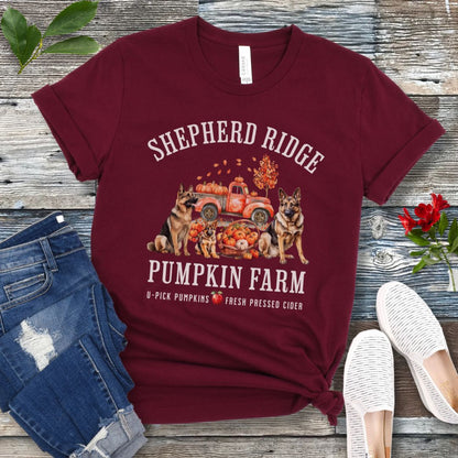 Maroon t-shirt with watercolor graphic of German Shepherds, a pumpkins truck and fall leaves, with the text 'Shepherd Ridge Pumpkin Farm', presented on a wooden surface with jeans and shoes.